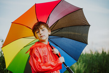 Portrait of a smiling school boy with rainbow umbrella in the park. Kid holds colourful umbrella on his shoulder. Cheerful child in a red raincoat holding multicolor umbrella outdoors.