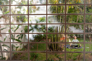 Woman practice yoga in the backyard surrounded by plants and trees, captured through the window of the house with tropical vibes and palm trees