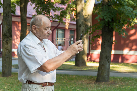 An elderly man walks alone in the park in the summer. A modern pensioner, businessman in a white shirt and trousers takes pictures with a camera in a mobile phone.