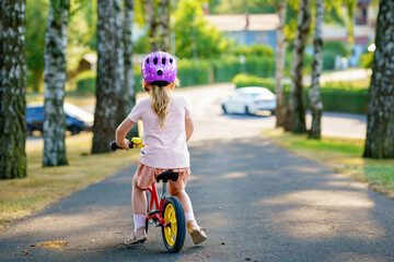 Little preschool girl with helmet running with balance bike on summer day. Happy child driving,...