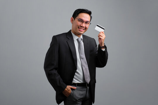 Portrait of smiling handsome young businessman in formal suit holding credit card isolated on grey background