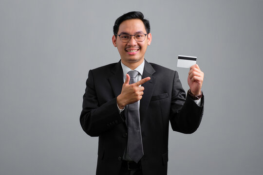 Portrait of smiling handsome young businessman in formal suit pointing at credit card isolated on grey background