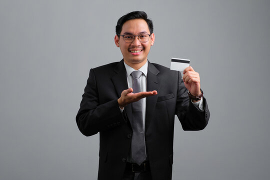 Portrait of smiling handsome young businessman in formal suit showing credit card isolated on grey background