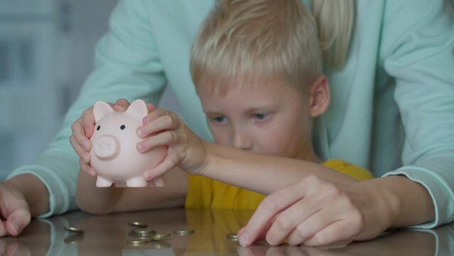 A boy of preschool age holds a piggy bank in the form of a pink pig in his hands. He shakes it with both hands and the coins he has been saving pour out