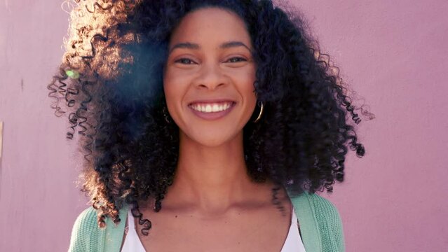 Portrait of a woman touching and moving her afro standing outdoor by a pink background. Happy, smile and beautiful latino girl embracing and playing with her natural curly hair while outside in sun.