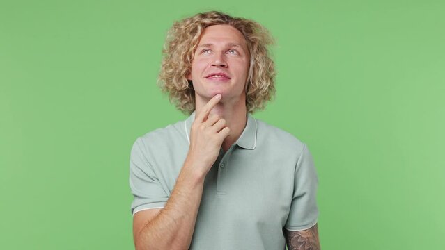Young fun caucasian blond man 20s he wear blue t-shirt look around think dream put hand prop up on chin lost in thought and conjectures isolated on plain pastel light green background studio portrait