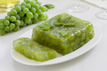 Sliced green grape jelly cake or white wine jelly on a white plate with bunch of grapes, glasses and bottle of wine on background. Selective focus, side view.
