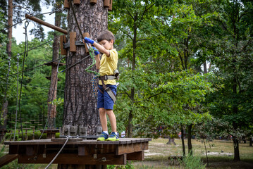 Strong excited young boy playing outdoors in rope park. Caucasian child dressed in casual clothes and sneakers at warm sunny day. Active leisure time with children concept