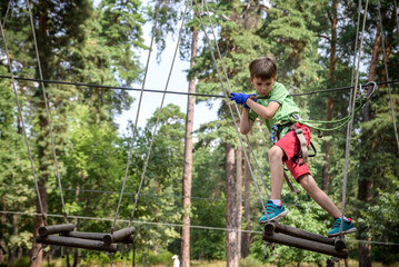 Obraz premium Strong excited young boy playing outdoors in rope park. Caucasian child dressed in casual clothes and sneakers at warm sunny day. Active leisure time with children concept