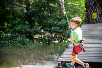 Strong excited young boy playing outdoors in rope park. Caucasian child dressed in casual clothes and sneakers at warm sunny day. Active leisure time with children concept