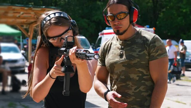 Strong long-haired caucasian girl pursuing her hobby by shooting from a black rifle with collimator. Male instructor in the background. Closeup portrait. Blurred foreground. . High quality 4k footage