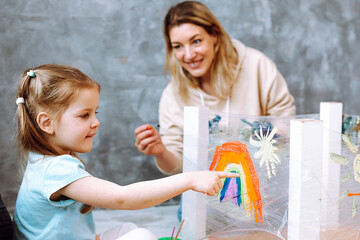 Female teacher with little blonde girl painting rainbow on oilcloth. Fun learning in kid development childcare center
