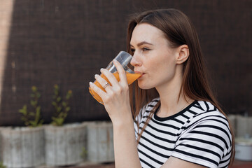 Young brunette, tranquil, relaxed brunette woman in striped t shirt drinking fresh organic orange juice in glass outdoor