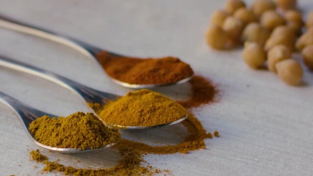 A spoonful of curry powder is placed on a table and some powder falls on the white tablecloth, nearby are two more spoons with turmeric powder and saffron powder and some chickpeas