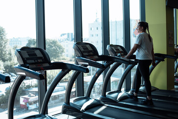 Plus size woman run on modern treadmill at fitness gym and look out window at city life. Middle...