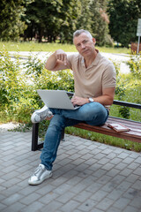 Aged gray haired businessman sitting on bench in sunny city park and pointing at digital laptop. Modern lifestyle, advertisement of remote job, outdoor workplace. Technology and people concept