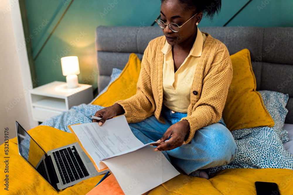 Wall mural young african american woman sitting in bed in the bedroom studying using laptop.