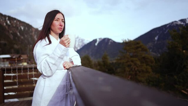 Young woman in a white coat, drinking morning coffee on terrace of hotel, looking at beautiful winter mountains. Woman drinking her morning coffee and enjoying view of mountains. Vacation lifestyle.
