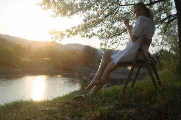 sideview of attractive blonde woman sitting on the chair raising her arms in rural lanscape against the riverside during the sunset