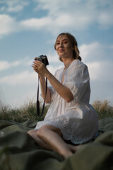 attractive caucasian woman with film camera in white sundress sitting on the meadow capturing the landscape against yhe blue sky