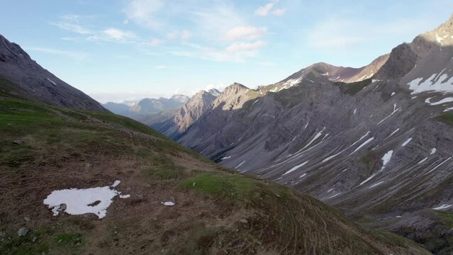 Aerial drone footage slowly rising upwards and looking across a dramatic, jagged mountain landscape with residual patches of snow and alpine meadows looking down a glacial valley in Switzerland.