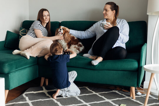Two tranquil and satisfied women and little boy sit on sofa, stroking dog Cavalier King Charles spaniel in living room