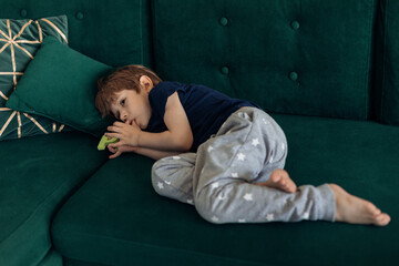 Portrait of upset, sad and depressed barefoot little boy lying on green sofa, sucking thumb finger....