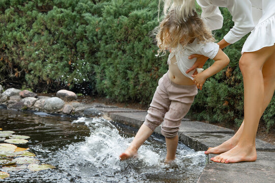 Unrecognizable woman holding blonde little girl, helping to wash legs in lake, pool water in natural park in hot summer