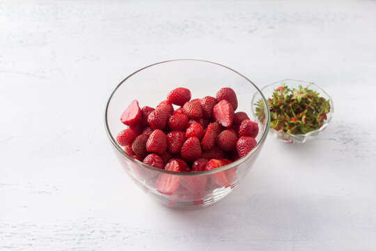 Glass bowl with peeled and chopped strawberries and a bowl with sepals on a light blue background