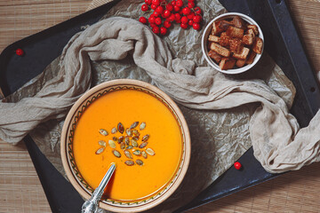 Cream yellow pumpkin soup squash with seeds, croutons near it, spoon in bowl on background of  baking paper, baking sheet, ash berry, napkin, autumn seasonal vegetarian healthy food, flatlay