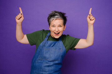 Excited attractive grey-haired woman happy and pointing by two hands and fingers above up isolated over violet background. Mature woman in apron playful pointing index by fingers above isolated