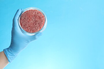 Scientist holding Petri dish with minced lab grown meat on light blue background, closeup. Space...