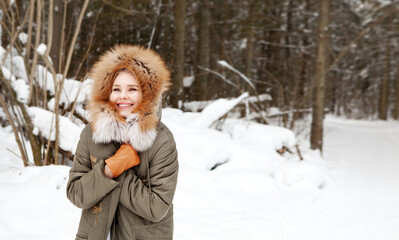 Happy woman smiles and wraps herself in warm clothes in winter forest on snow background. Wearing stylish fashion outfit khaki parka coat jacket with fur hood