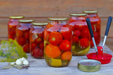 glass jars with canned tomatoes with iron lids stand on a wooden table with garlic, dill, iron lids and a seaming machine. home storage solution, saving leftovers, canning concept, rustic composition