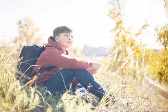 Autumn portrait of teenager boy sitting on grass. Young man with backpack resting relaxing, contemplating, thinking, daydreaming. Teen deep in thought Local travel. Active lifestyle People from behind