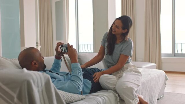 Love, photography and man taking a picture of his wife on a camera while relaxing on a sofa together. Happy, smile and woman showing peace sign for a photo while sitting on the couch with her husband