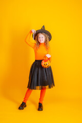 a teenage girl in a witch costume on a yellow background, holding a confent pumpkin eating marmalade worms halloween party