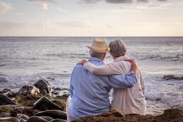 Rear view of relaxed caucasian senior couple sitting on the pebble beach at sunset light admiring...