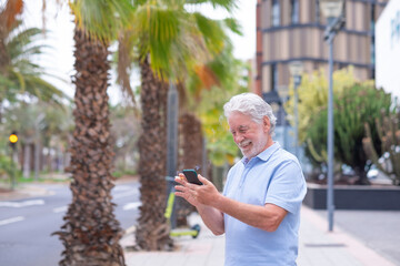 Smiling bearded caucasian senior man using mobile phone standing outdoor in the city street, smoking a cigarette. Palm trees on background