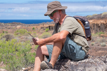 Senior bearded man with hat and backpack sitting on the rocks resting after trekking in countryside - horizon over sea in background