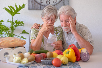 Portrait of sad, unhappy, worried, old caucasian couple sitting at home table looking at grocery...
