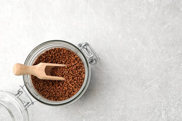 Glass jar with wooden scoop and buckwheat tea granules on light grey table, top view. Space for text