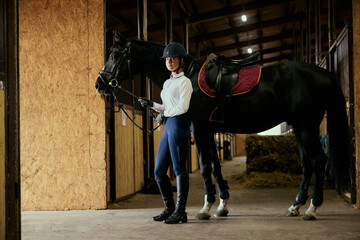 Live portrait of young girl, teen, beginner rider in sports uniform and helmet going with horse to arena. Lifestyle mood. Concept of nature, animal, care, sport