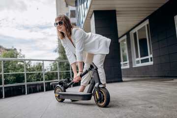 Pretty woman in white suit folding her electro scooter after ride while standing on background of modern building