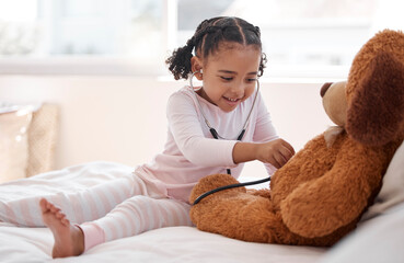 A black girl playing hospital doctor with a teddy bear on her bed, holding a stethoscope and...