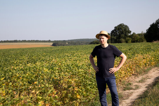 A farmer in a straw hat and dark blue clothes looks on agricultural field with soybeans. Front view
