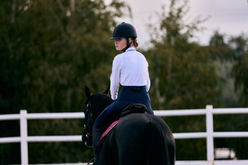 Sports training. Young sportive girl, professional rider in sports uniform and helmet practicing at riding arena in summer day, outdoors. Horseback riding