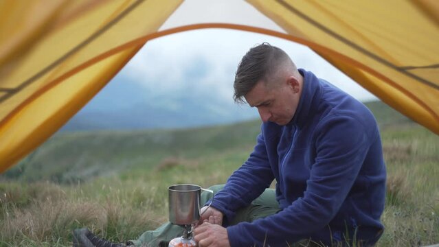 A guy cooks food on a gas burner in the mountains. A tourist on a halt during a hike in the mountains with a tent heats water in a cup. Concept of tourism in a landscape place. High quality 4k footage