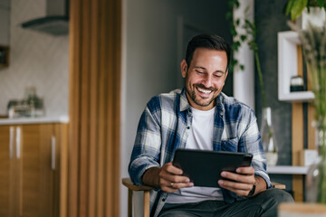 Photo of a happy man holding and using a digital tablet, sitting on the armchair.