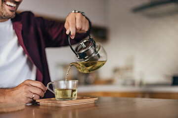Photo of a smiling man sitting in the kitchen, pouring tea in a cup.